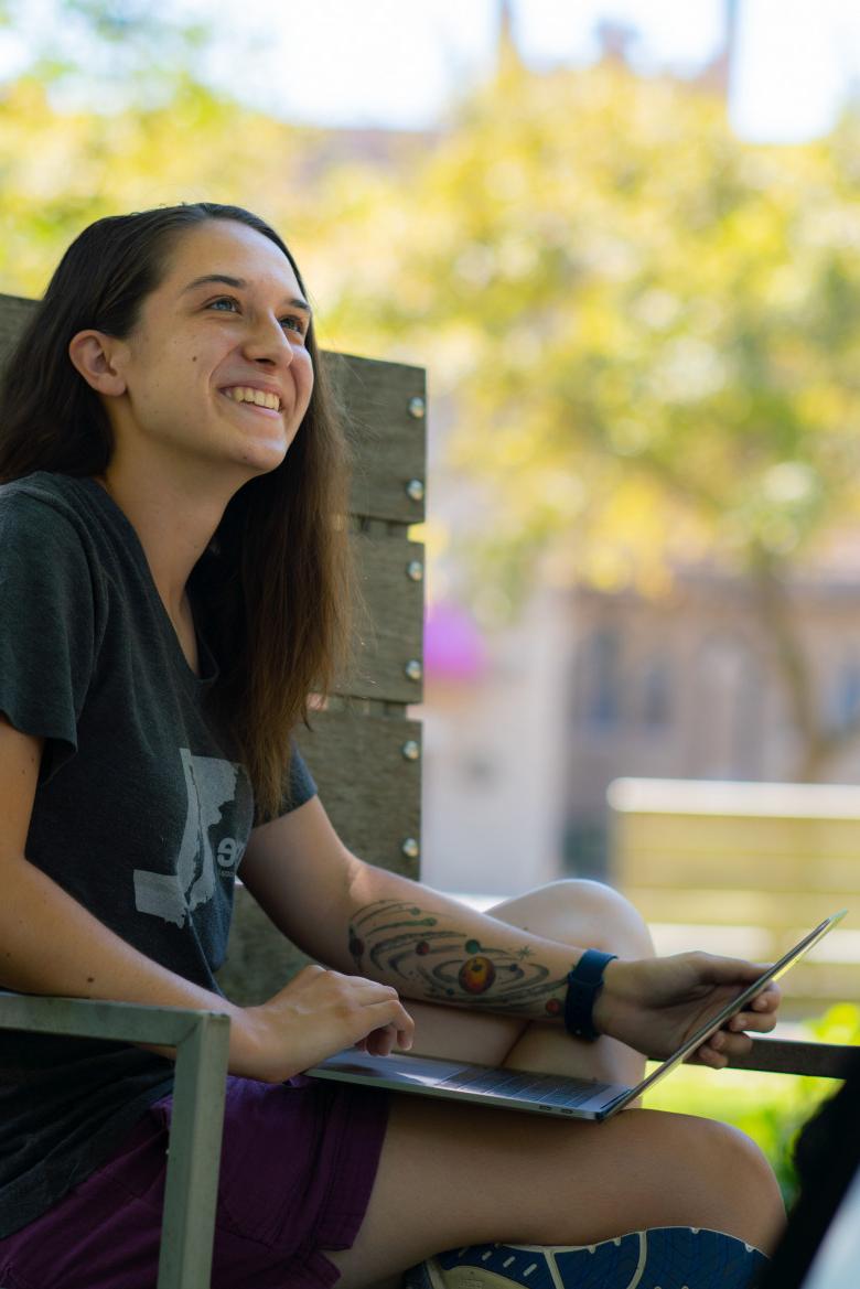 student sits on a bench in the poetry garden with a laptop and green foliage around